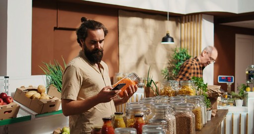 Man with a beard holding a smartphone in a kitchen setting with jars and produce.